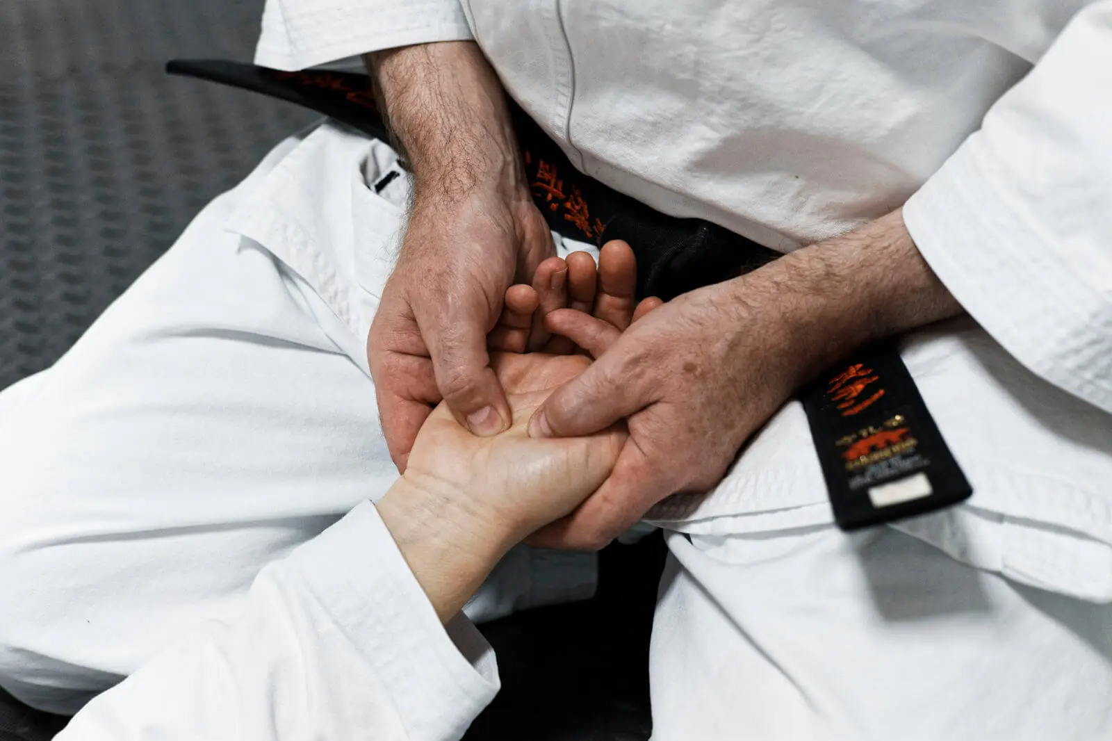 Ein sitzender Mann im Karateanzug drückt mit beiden Daumen auf der Handfläche einer weiblichen Hand