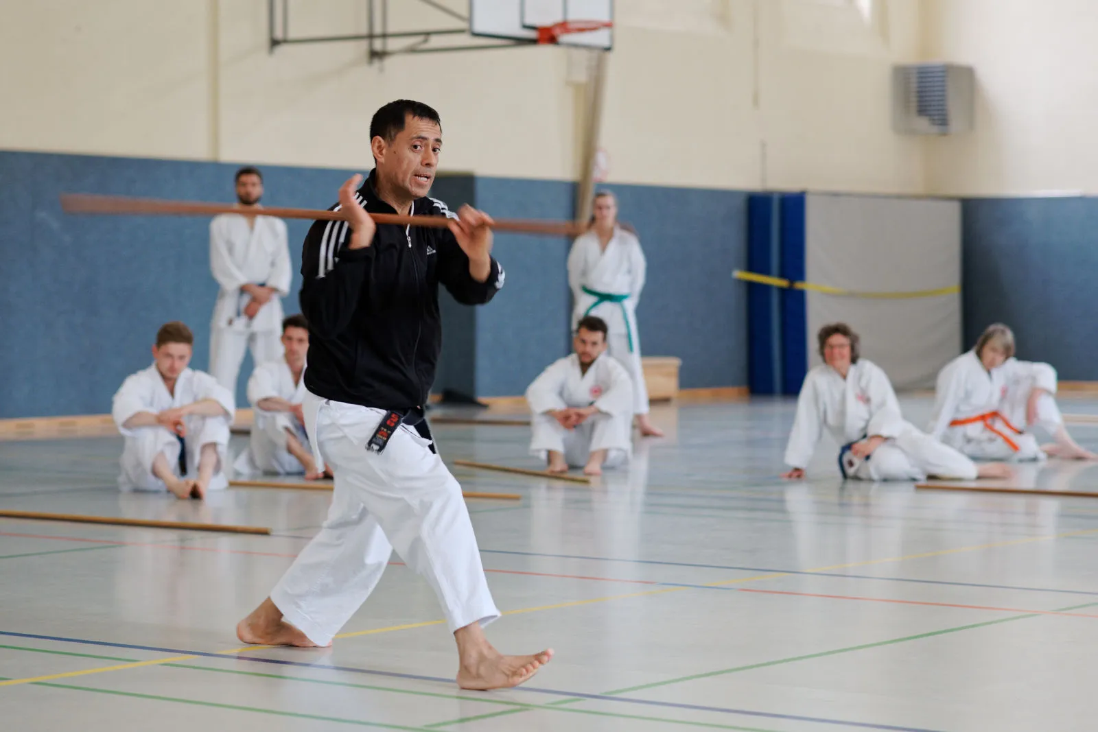 Roberto Romero Sensei schlägt mit einem Bo in einer Sporthalle mit mehreren Personen im Hintergrund