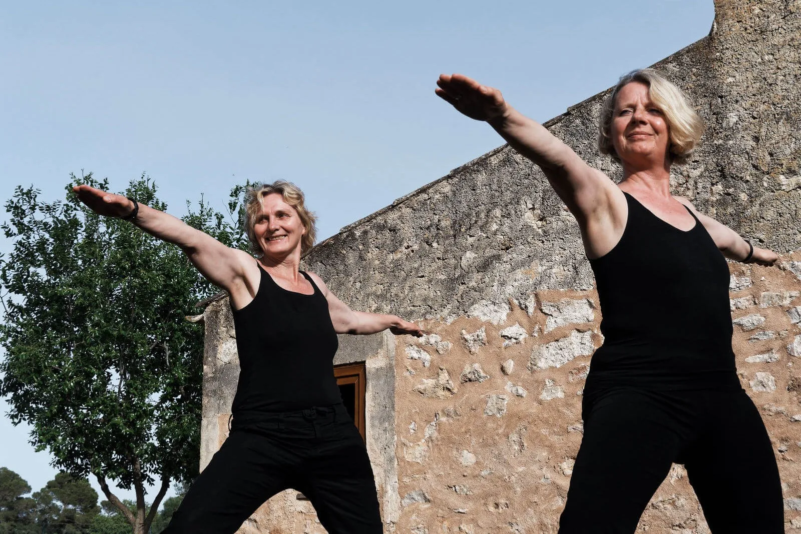 Zwei Frauen in schwarzer Kleidung stehen vor einer Steinmauer in der Yoga-Kriegerpose