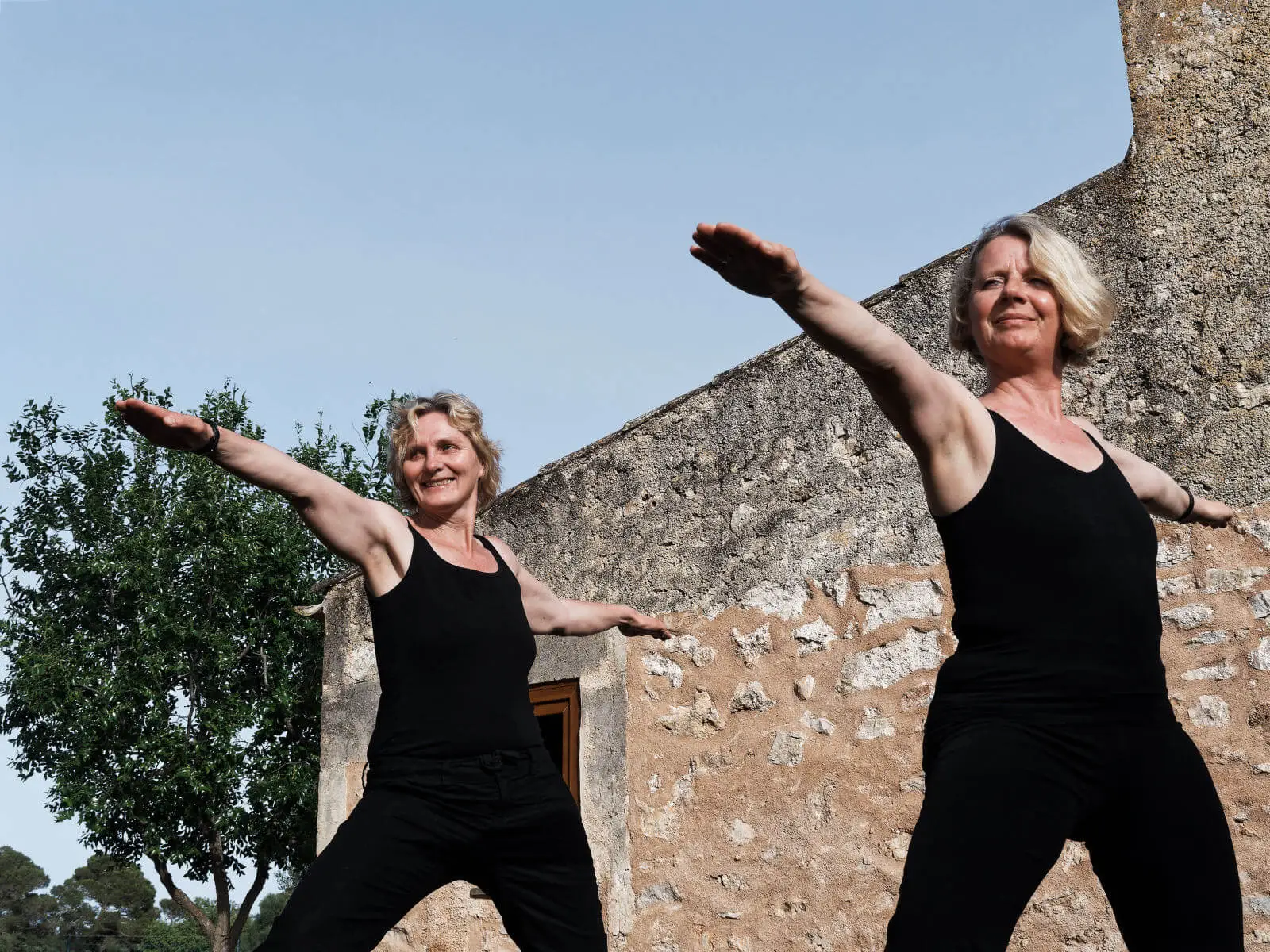 Zwei Frauen in schwarzer Kleidung stehen vor einer Steinmauer in der Yoga-Kriegerpose